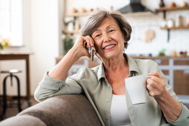 Cheerful caucasian senior old woman talking on smart phone with relatives doctor while drinking